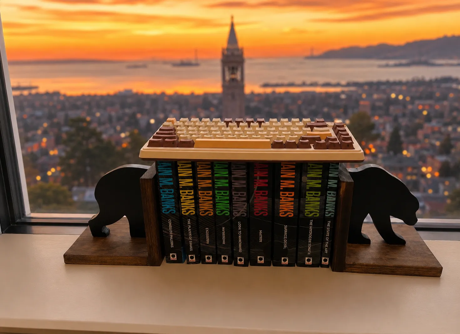 A keyboard beside a shelf of Iain M. Banks' Culture novels on a windowsill, overlooking UC Berkeley's Sather Tower at sunset.